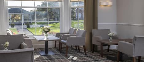 A room with a dining table, four chairs, a small side table, and a view of a garden through large windows.