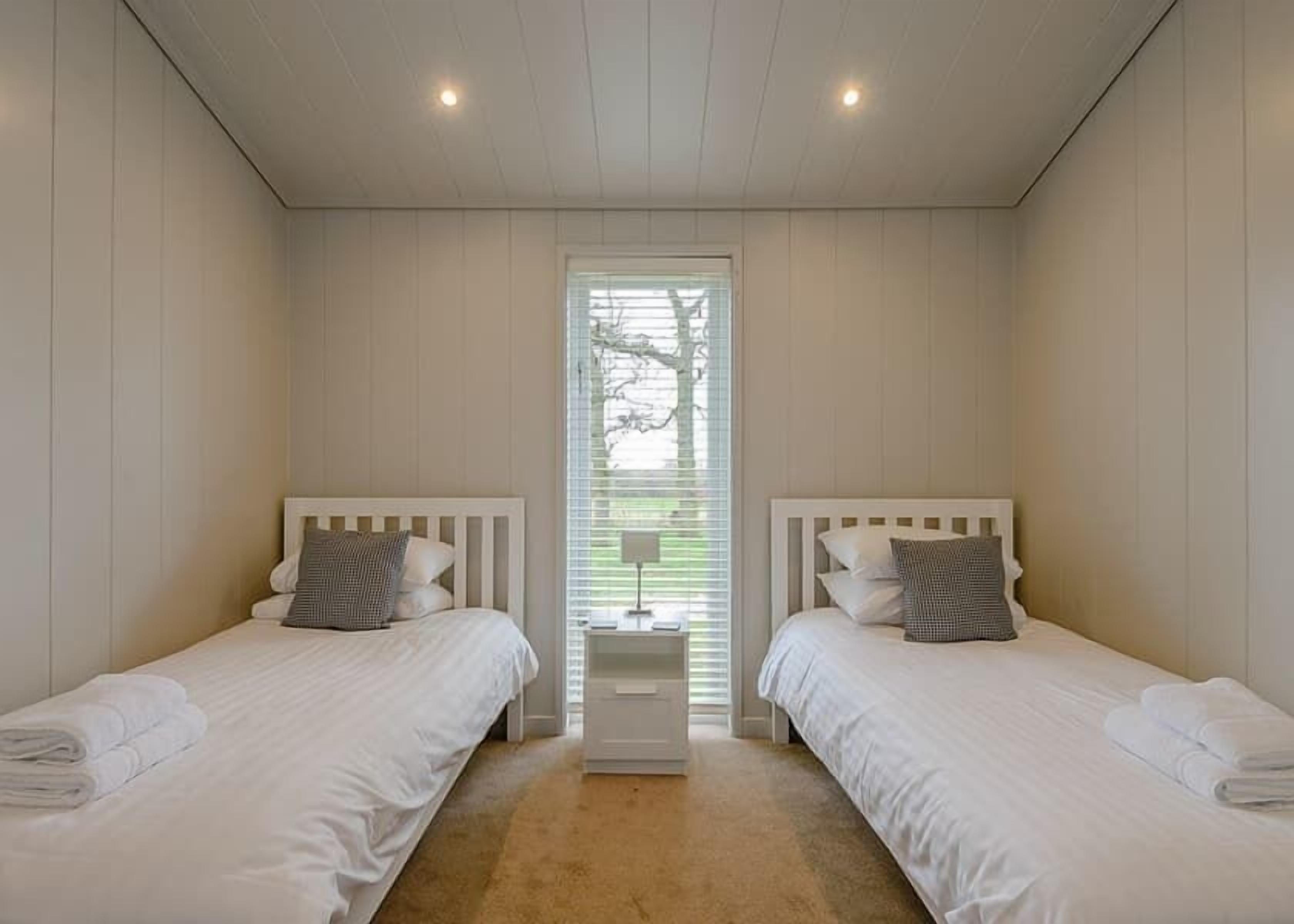 A bedroom with two beds, a window with blinds, and a nightstand with a lamp.