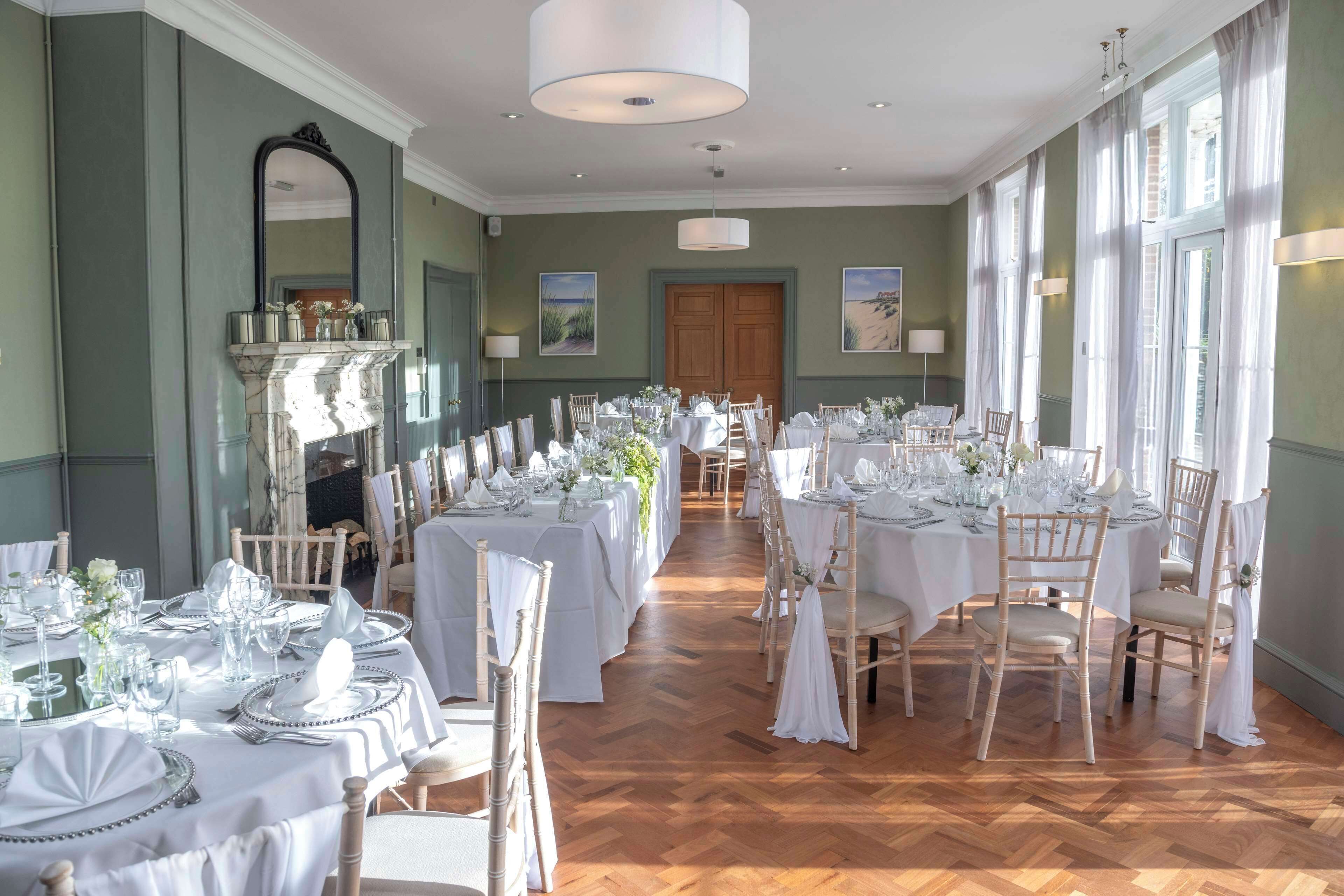 A formal dining room with multiple tables set for a banquet, featuring white tablecloths, glassware, and wooden chairs.