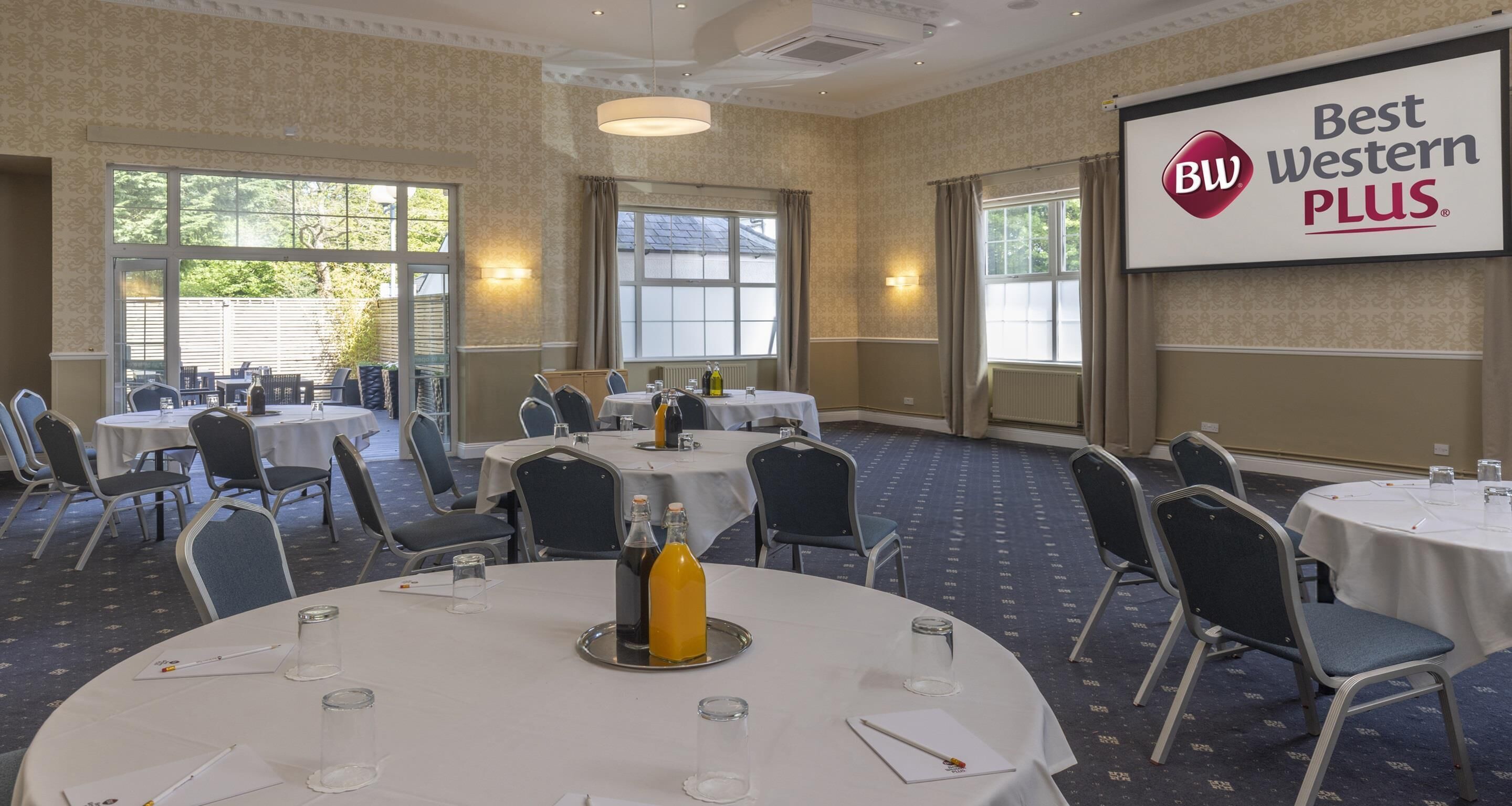 Een congreszaal met ronde tafels, stoelen en een projectiescherm waarop 'Best Western PLUS' staat.