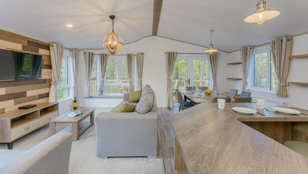 Een moderne woonkamer met een bank, salontafel en een aan een houten wand bevestigde televisie. Er zijn ramen met gordijnen, een eethoek met tafel en stoelen, en een keuken met een aanrecht en krukjes.