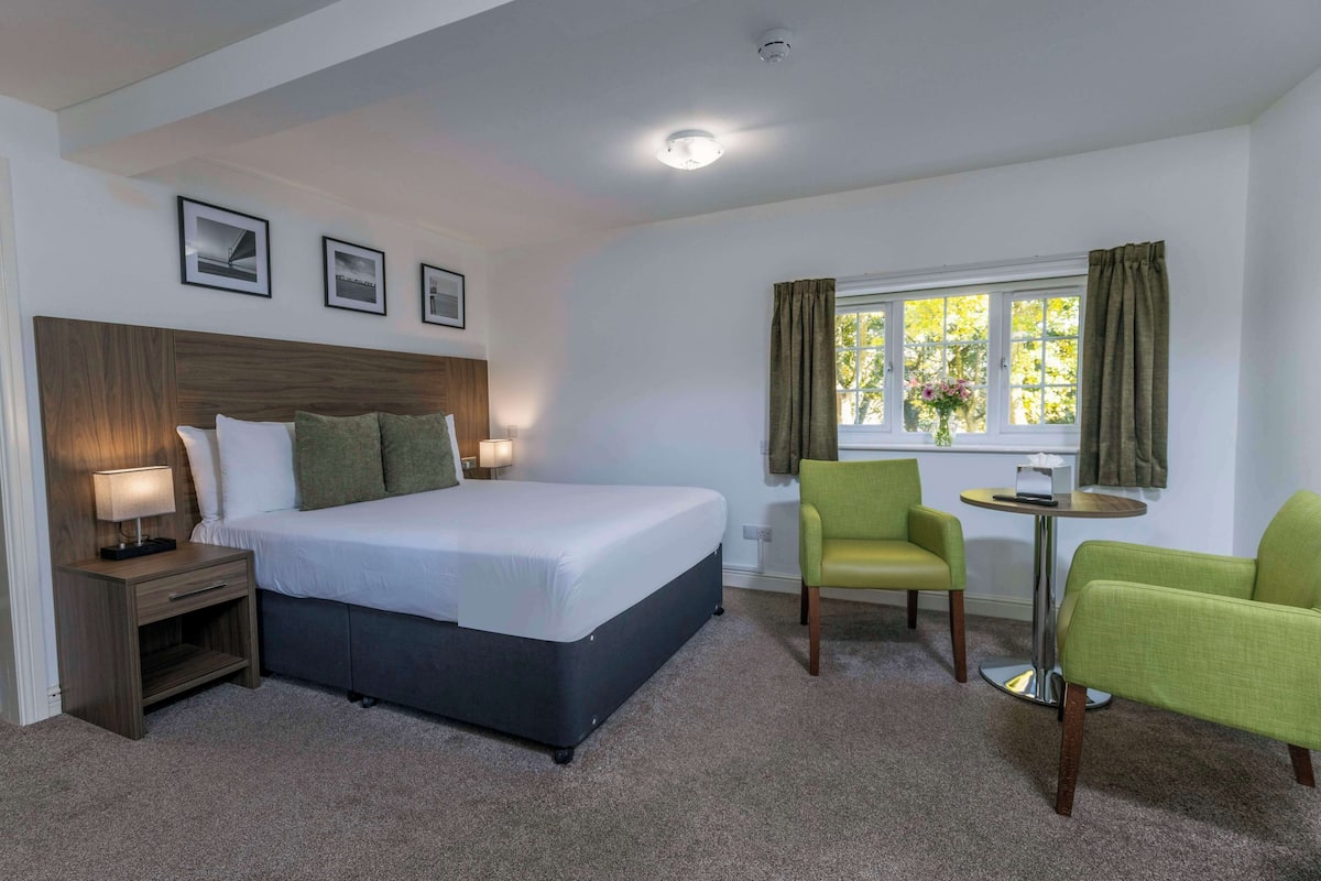 Een hotelkamer met een bed, nachtkastje, stoel, een klein tafeltje en een raam met gordijnen.