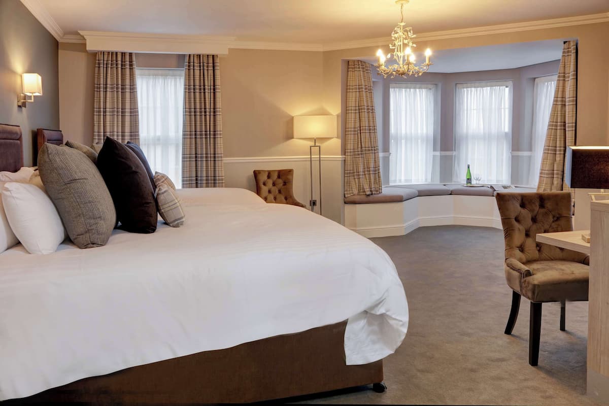 Een groot bed met veel kussens, een nachtkastje, een stoel, een lamp en uitzicht op een raam met gordijnen.