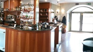 A bar area with a wooden counter, bar stools, and a variety of bottles and glasses. There is a large window with a view of the outside. - Azalea Hotel (Baveno)