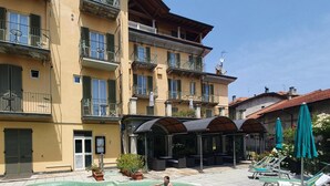 A swimming pool surrounded by a courtyard with lounge chairs and umbrellas, a covered seating area, and multi-story buildings with balconies. - Azalea Hotel (Baveno)