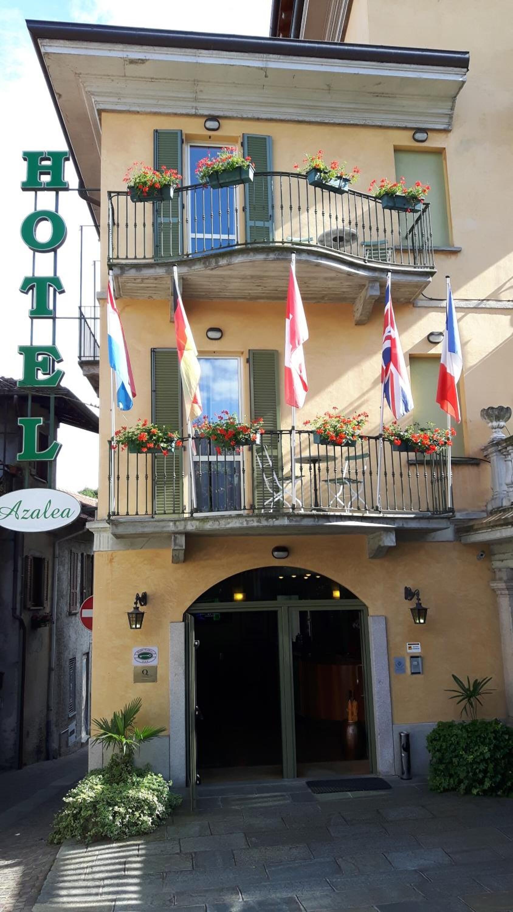 Hotellin edessä on kyltti, jossa lukee ”HOTEL Azalea”. Rakennus on kaksikerroksinen, ja sen parvekkeet on koristeltu kukilla täytetyillä kukkaruukuilla.