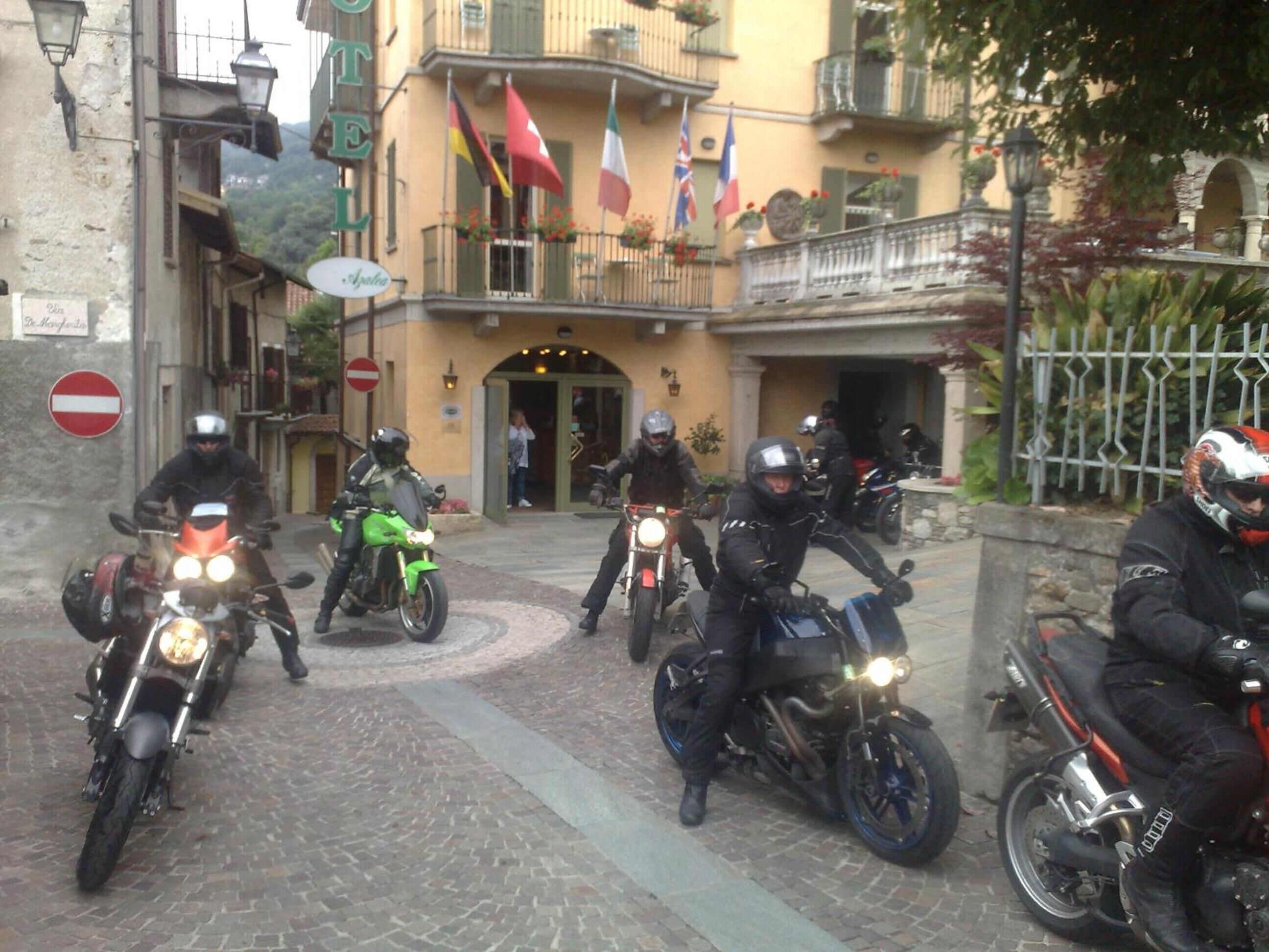 Un grupo de motociclistas estacionados frente a un edificio con varias banderas, una calle adoquinada y una señal de 'prohibido el paso'.