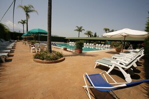 Un espace aménagé au bord de la piscine, avec des chaises longues et des parasols, une piscine et des palmiers.