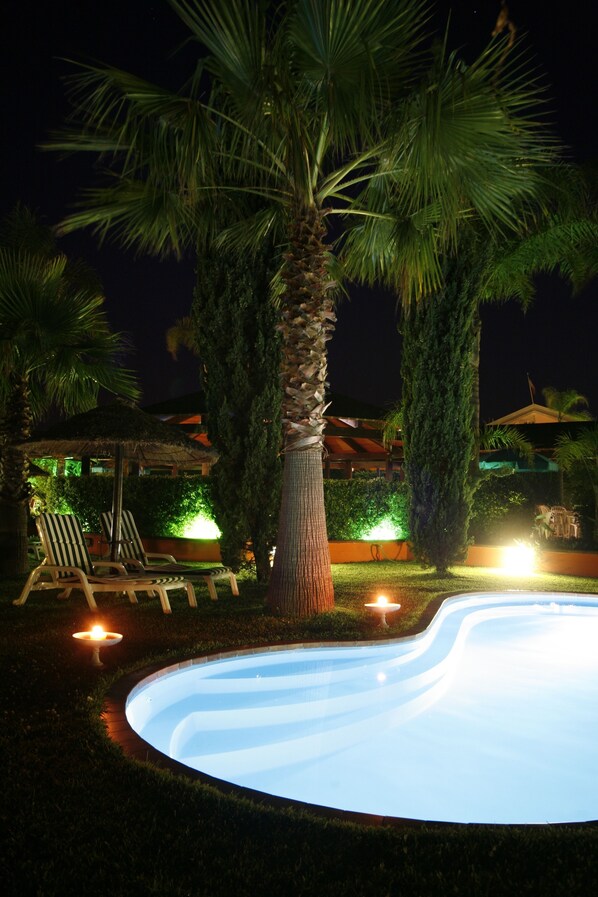 Una piscina rodeada de palmeras y sillas de descanso al caer la noche.