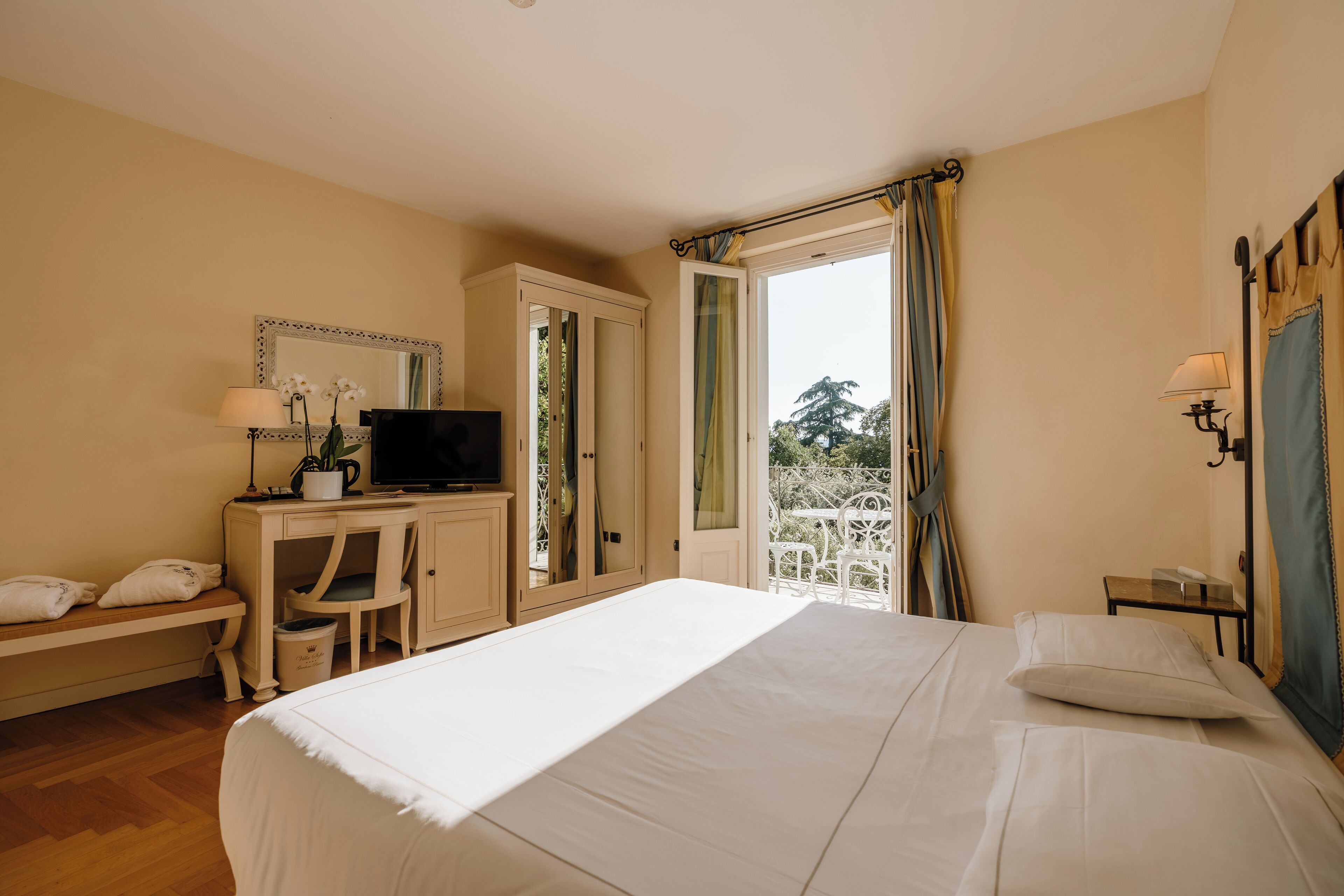 A bedroom with a bed, a desk with a television, a chair, a bench, and a window with curtains.