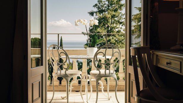 An open balcony with two white metal chairs and a table, overlooking a scenic view of trees and the sea. - Hotel Villa Sofia (Gardone Riviera)