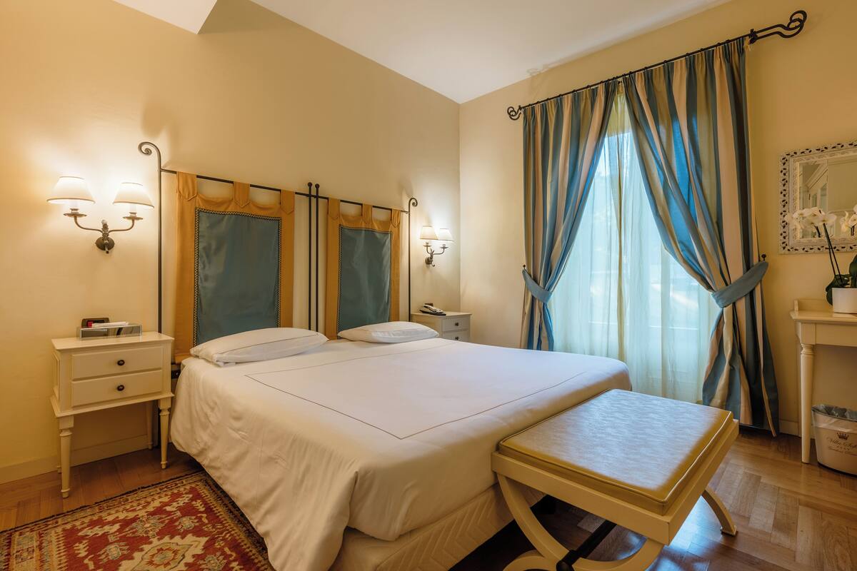 Une chambre à coucher bien rangée, avec un lit, des tables de chevet, des lampes et une fenêtre avec des rideaux.