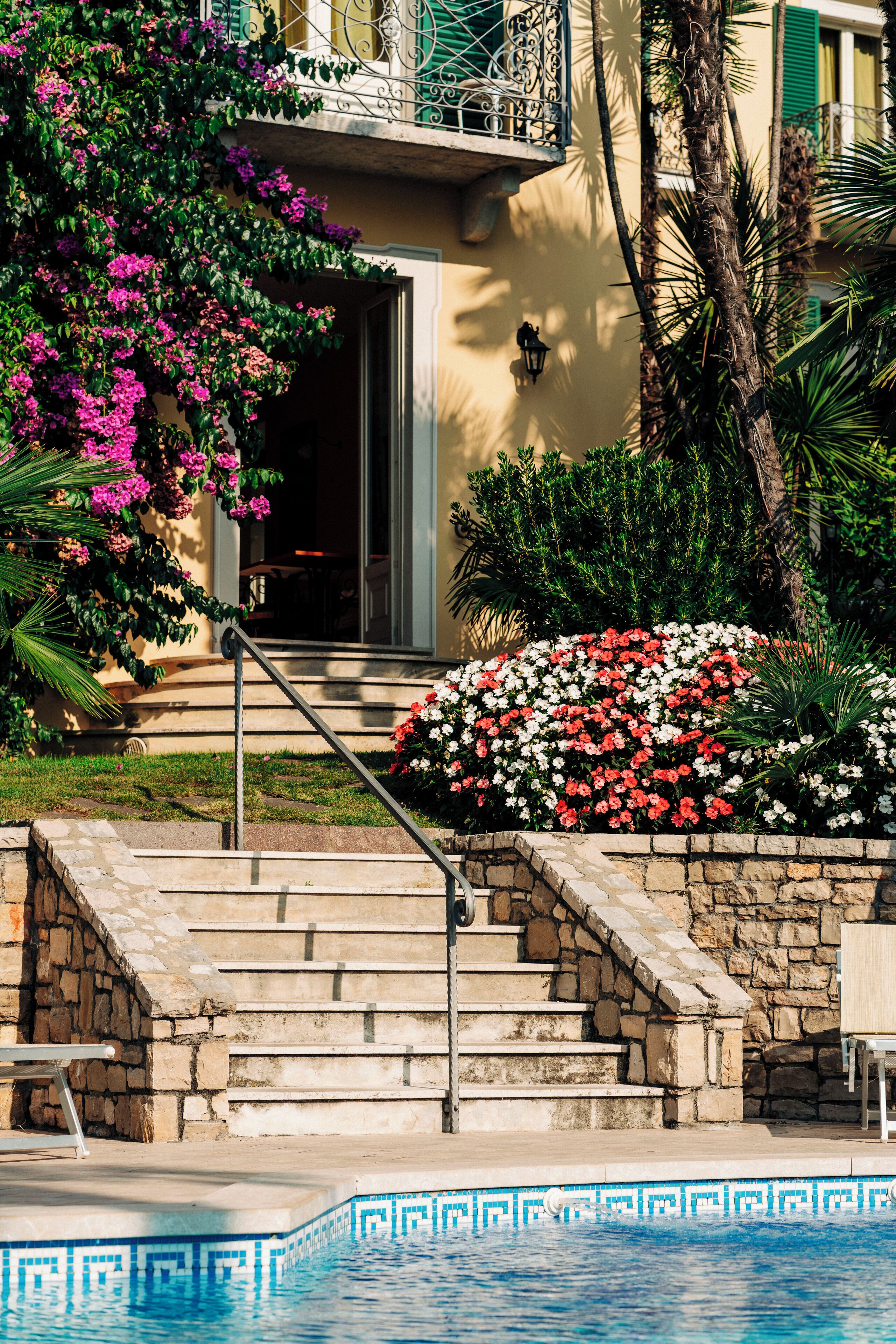 Una zona de piscina con escalones de piedra que conducen a la entrada de un edificio, rodeada de flores y follaje vibrantes.