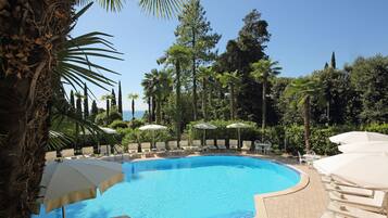 Piscina con sillones de playa y sombrillas, rodeada de palmeras y otra vegetación.