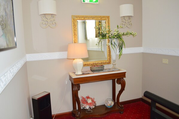 A hallway with a mirror, a lamp, a vase with flowers, a small table, and a red carpet.