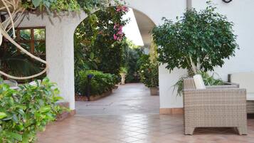 Un espacio al aire libre techado, con suelo de baldosas, una silla de mimbre y varias plantas en macetas.