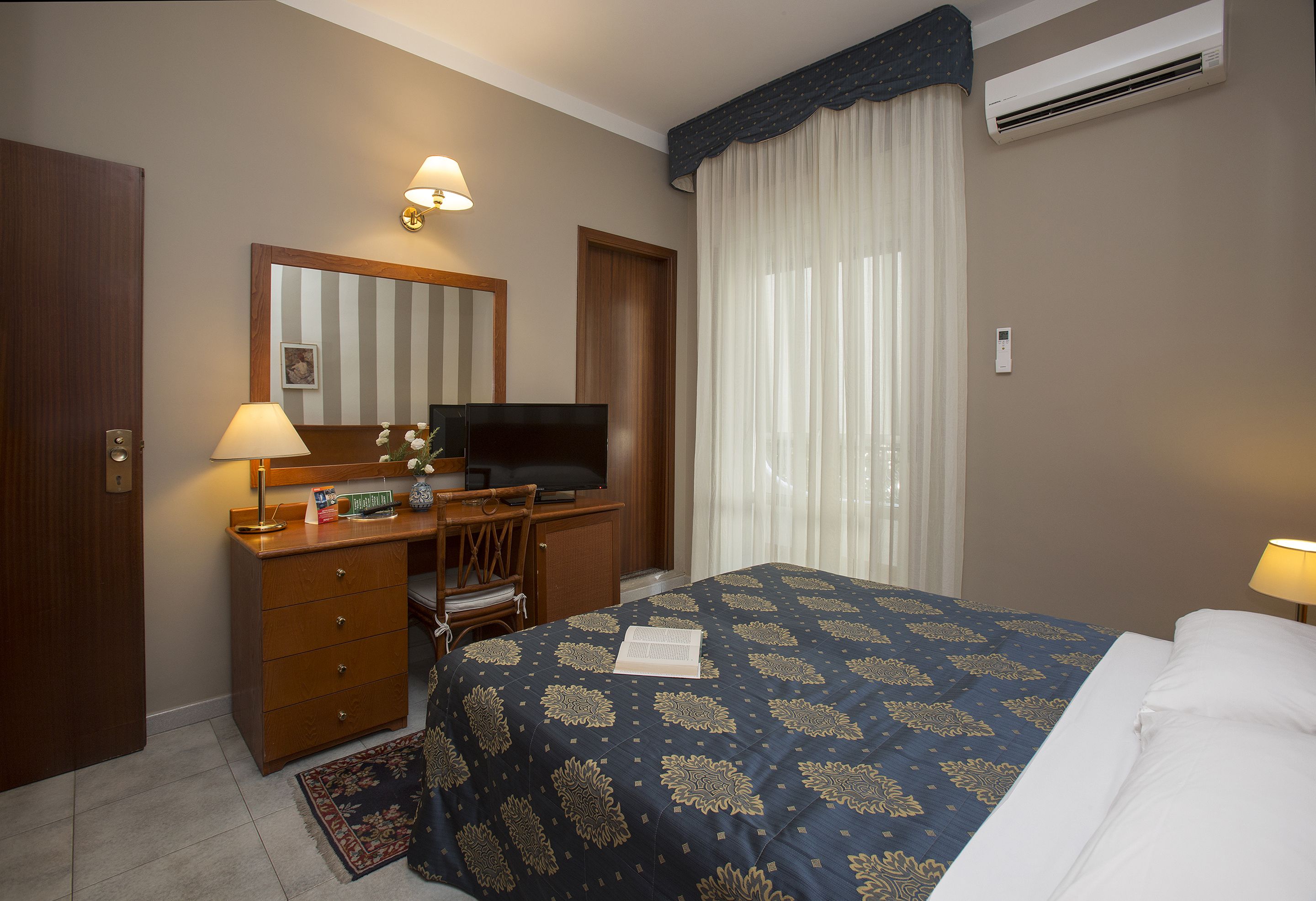 Habitación de hotel con cama, escritorio con televisor, cómoda y ventana con cortinas.