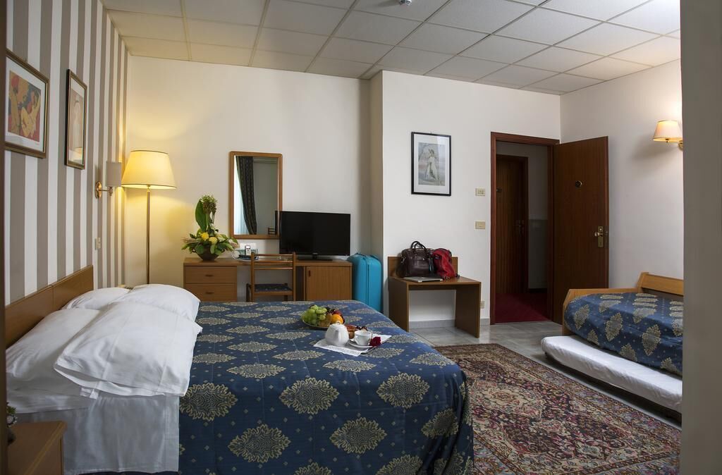 Habitación de hotel con cama, escritorio, televisor, silla, lámpara de escritorio, espejo, puerta y una alfombra pequeña.