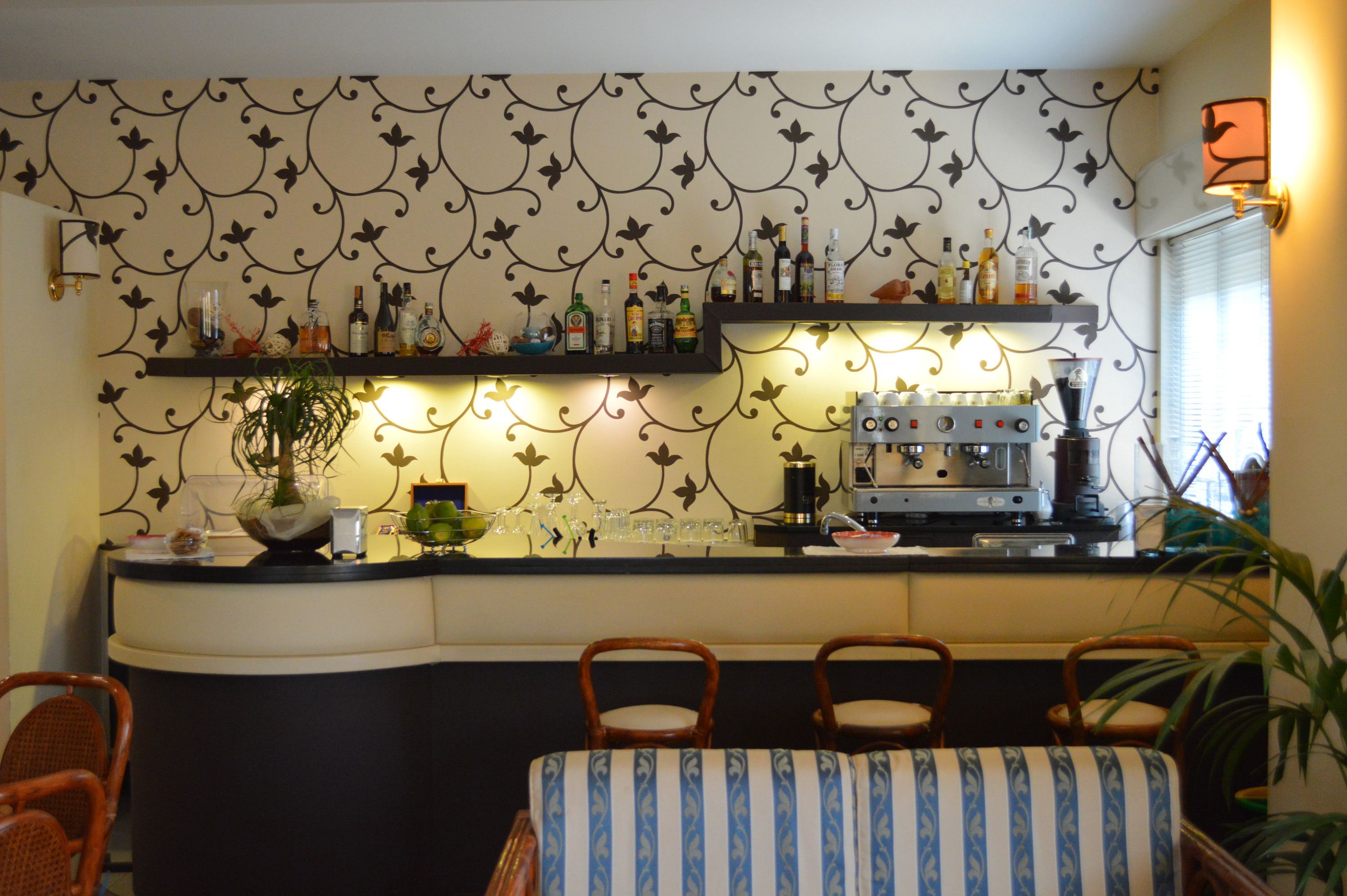 Un coin bar avec des étagères remplies de bouteilles, un appareil à café et une plante en pot. On peut voir des chaises et une table au premier plan.