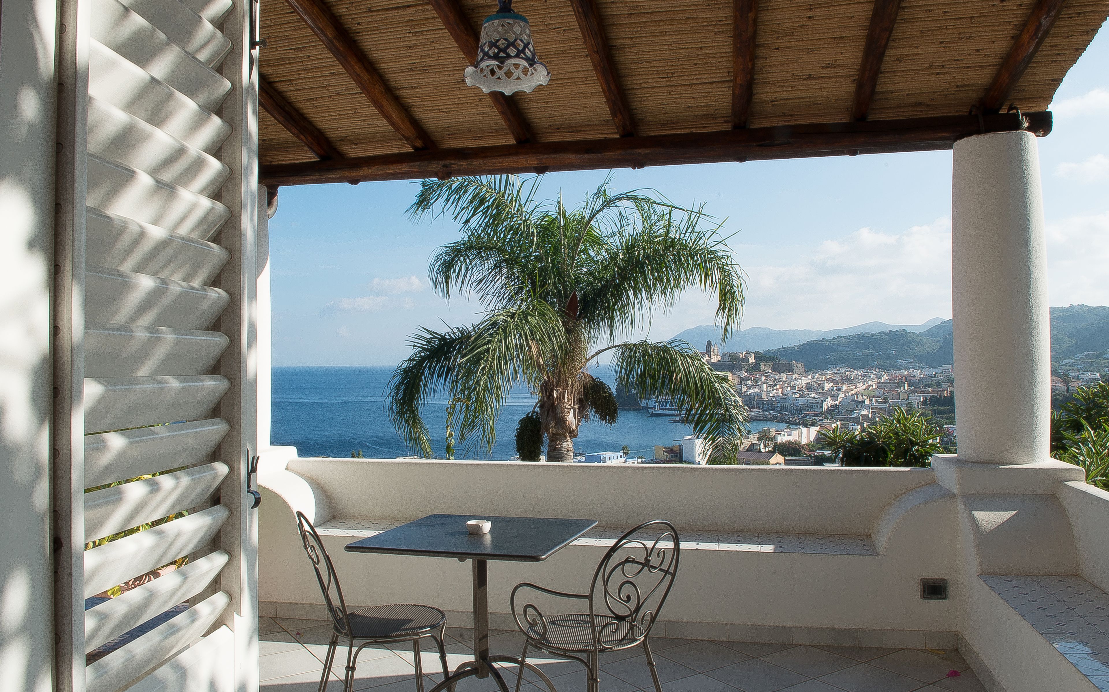 A covered outdoor seating area with a table and chairs, overlooking a coastal cityscape with a palm tree and mountains in the distance.