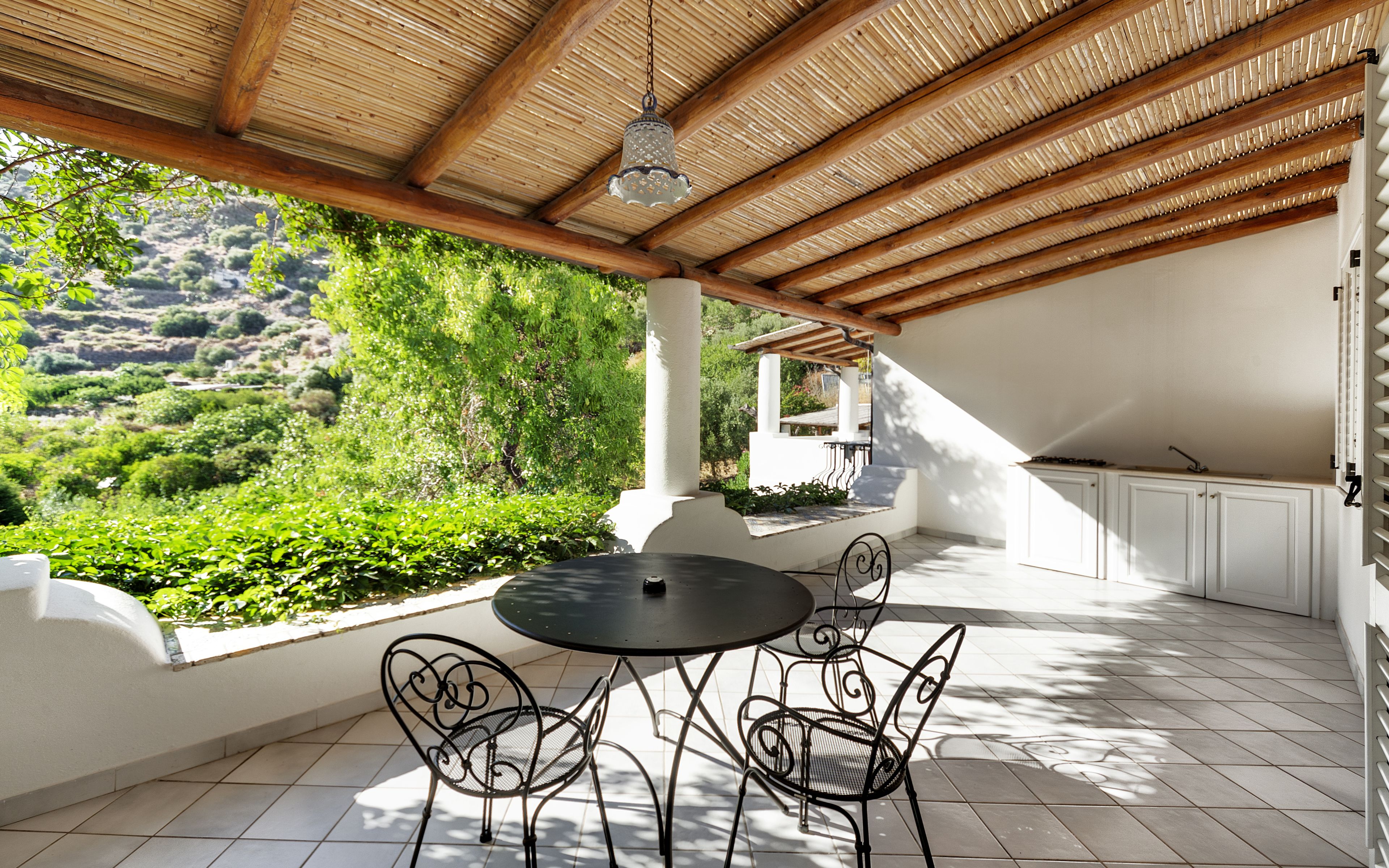 A covered patio with a table and chairs, a view of greenery, and a kitchen area.