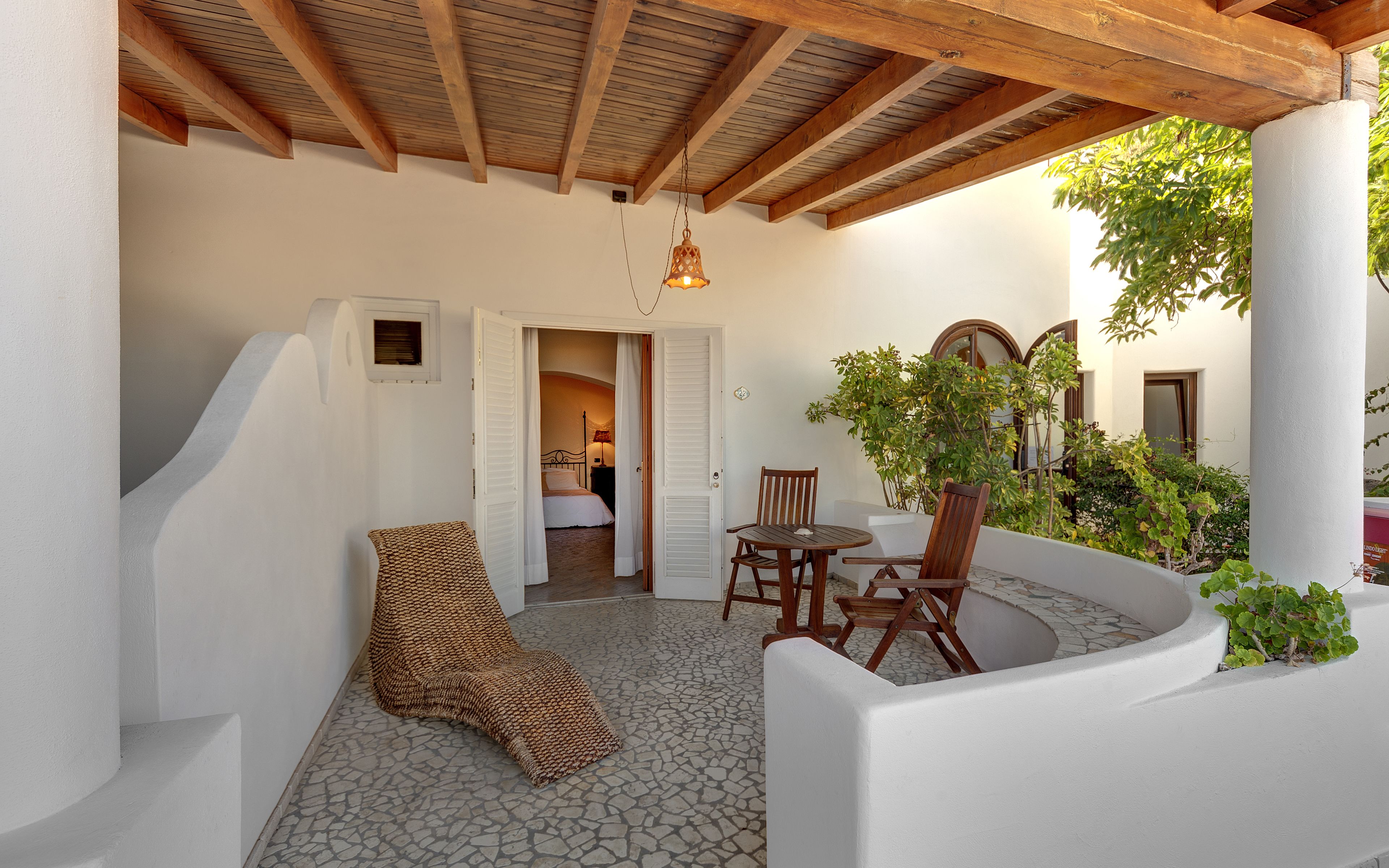 A cozy patio with a wooden ceiling, a woven chair, a table, and a potted plant.