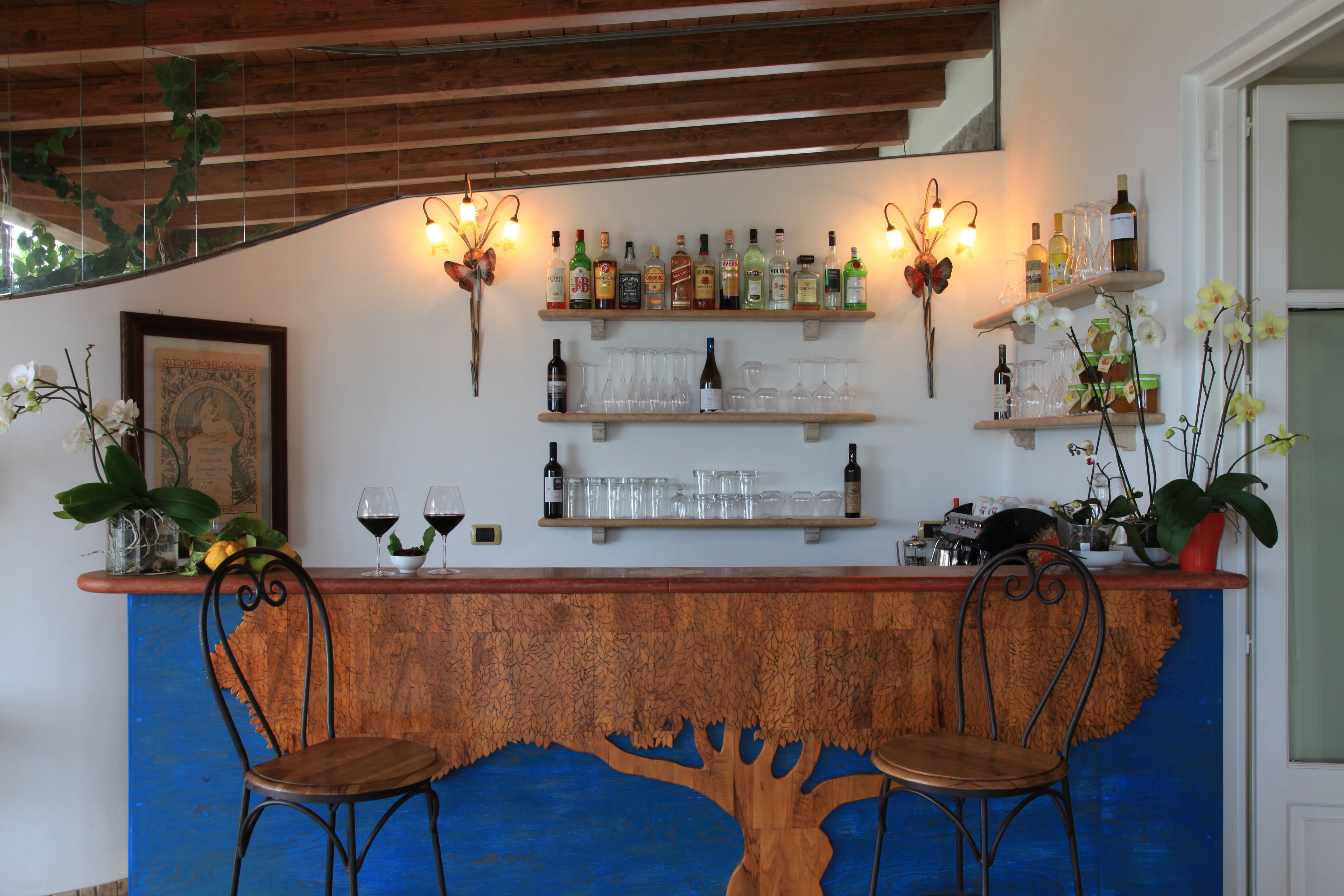 A bar area with wooden shelves displaying bottles and glasses, two chairs, and decorative plants.