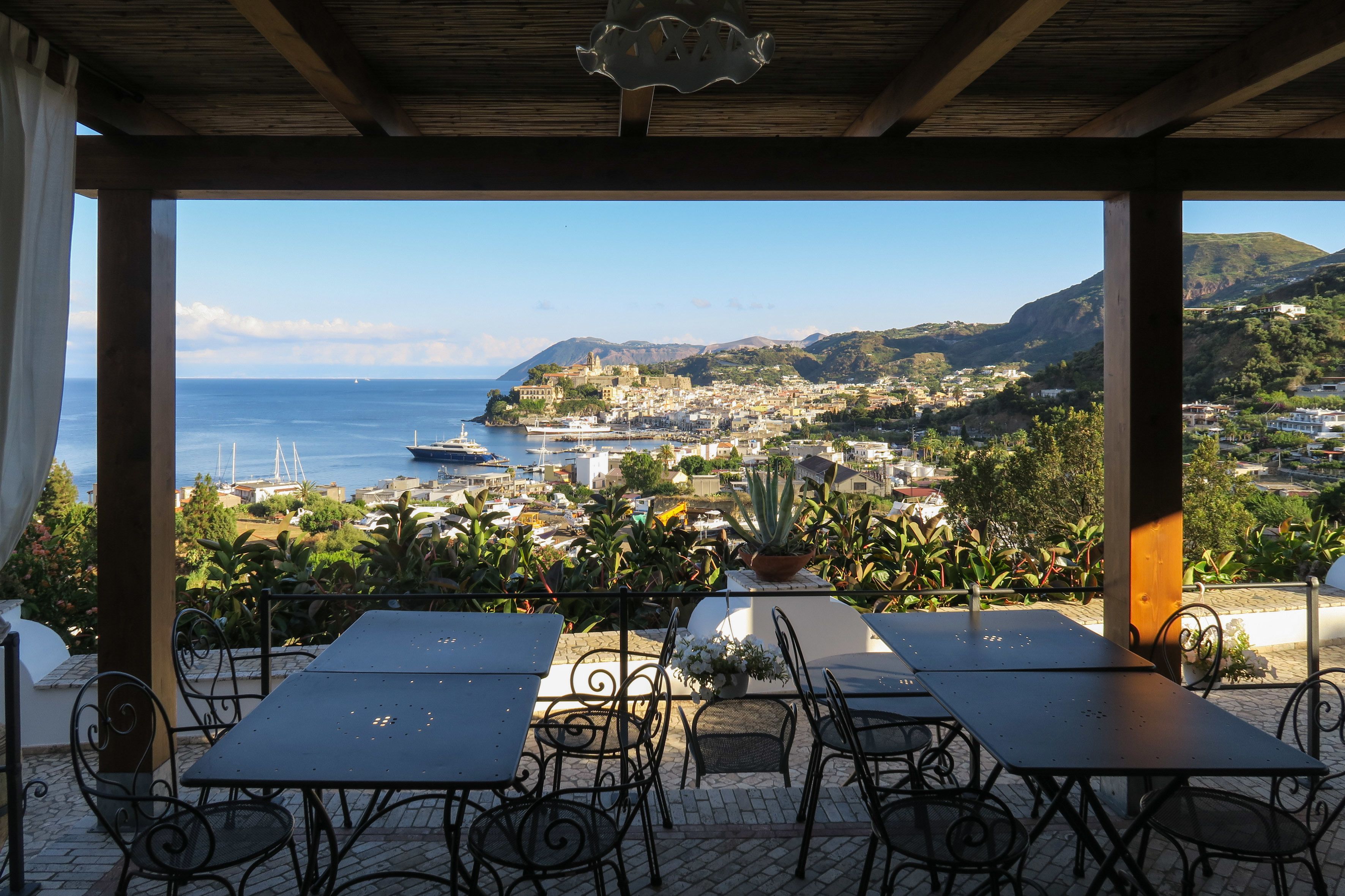 An outdoor patio with tables and chairs, overlooking a coastal town with buildings and boats.