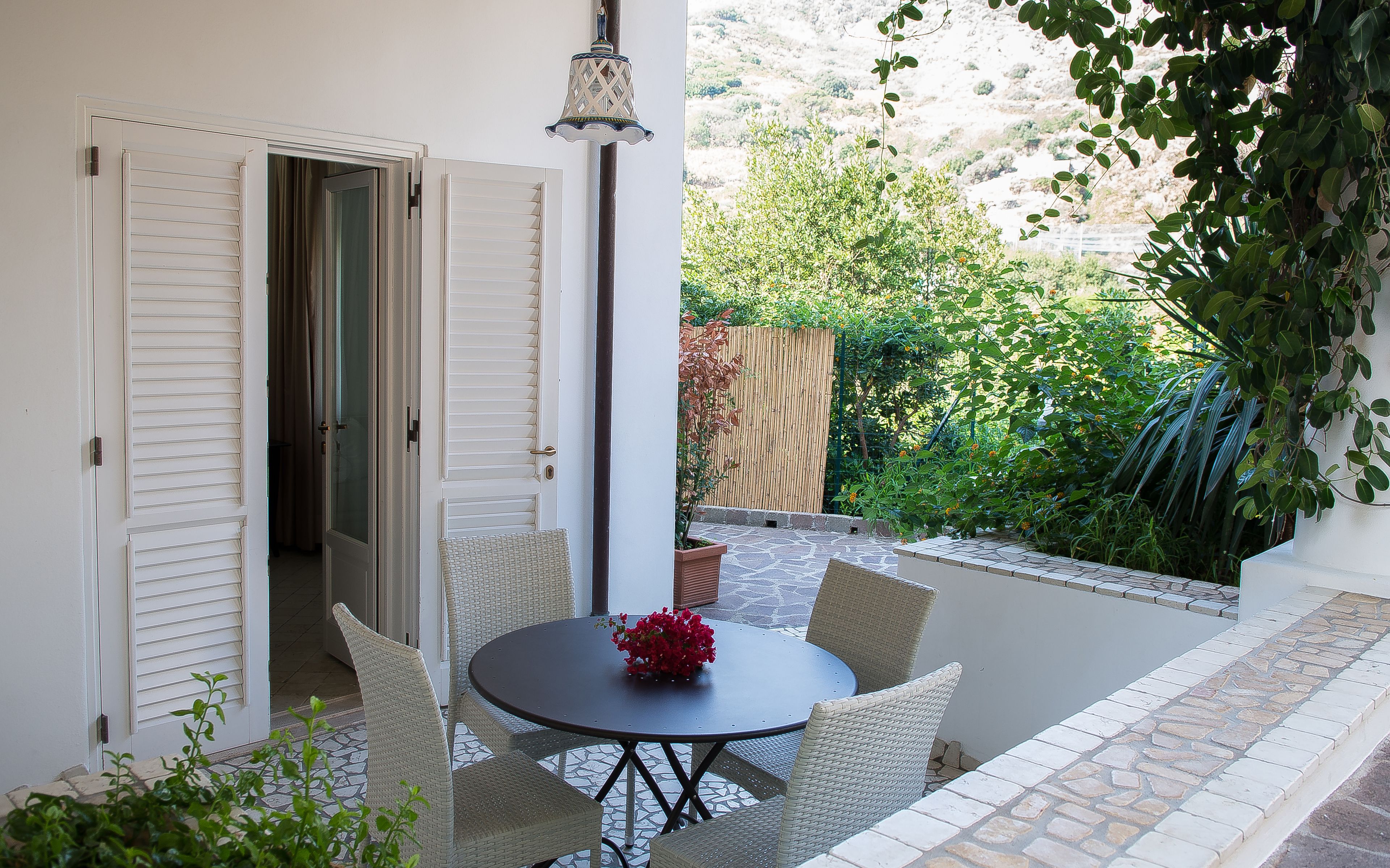 Un patio al aire libre con una mesa redonda y sillas de mimbre, una planta en maceta y vistas de un jardín con cerca de madera y follaje.