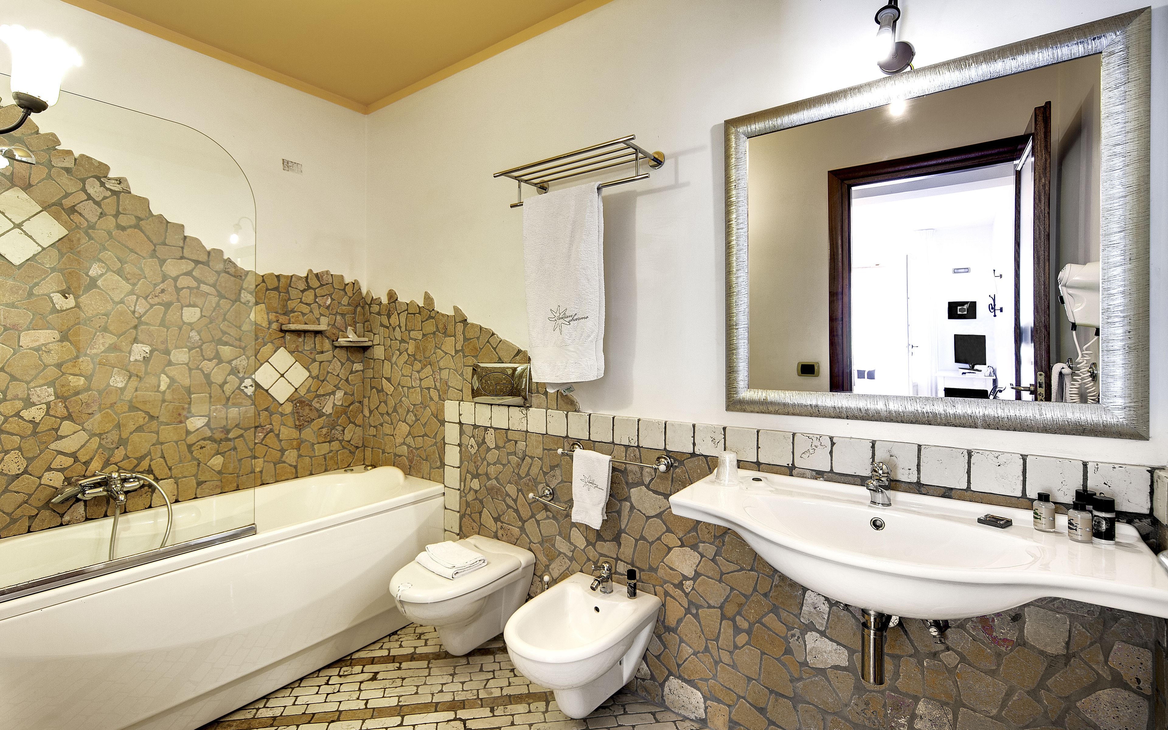 A bathroom with a bathtub, bidet, and sink, featuring a stone mosaic wall and a large mirror.