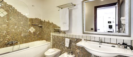 A bathroom with a bathtub, bidet, and sink, featuring a stone mosaic wall and a large mirror.