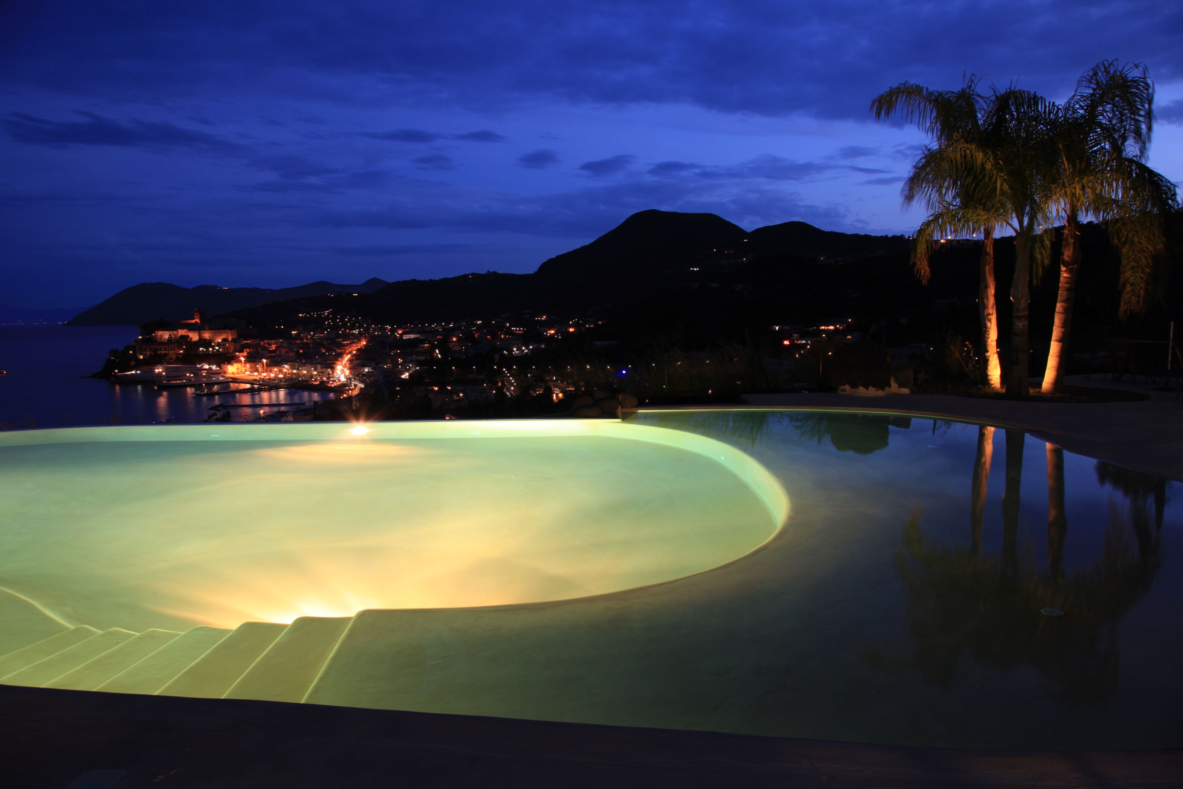 A swimming pool with a curved edge, surrounded by palm trees and a view of a coastal town at night.
