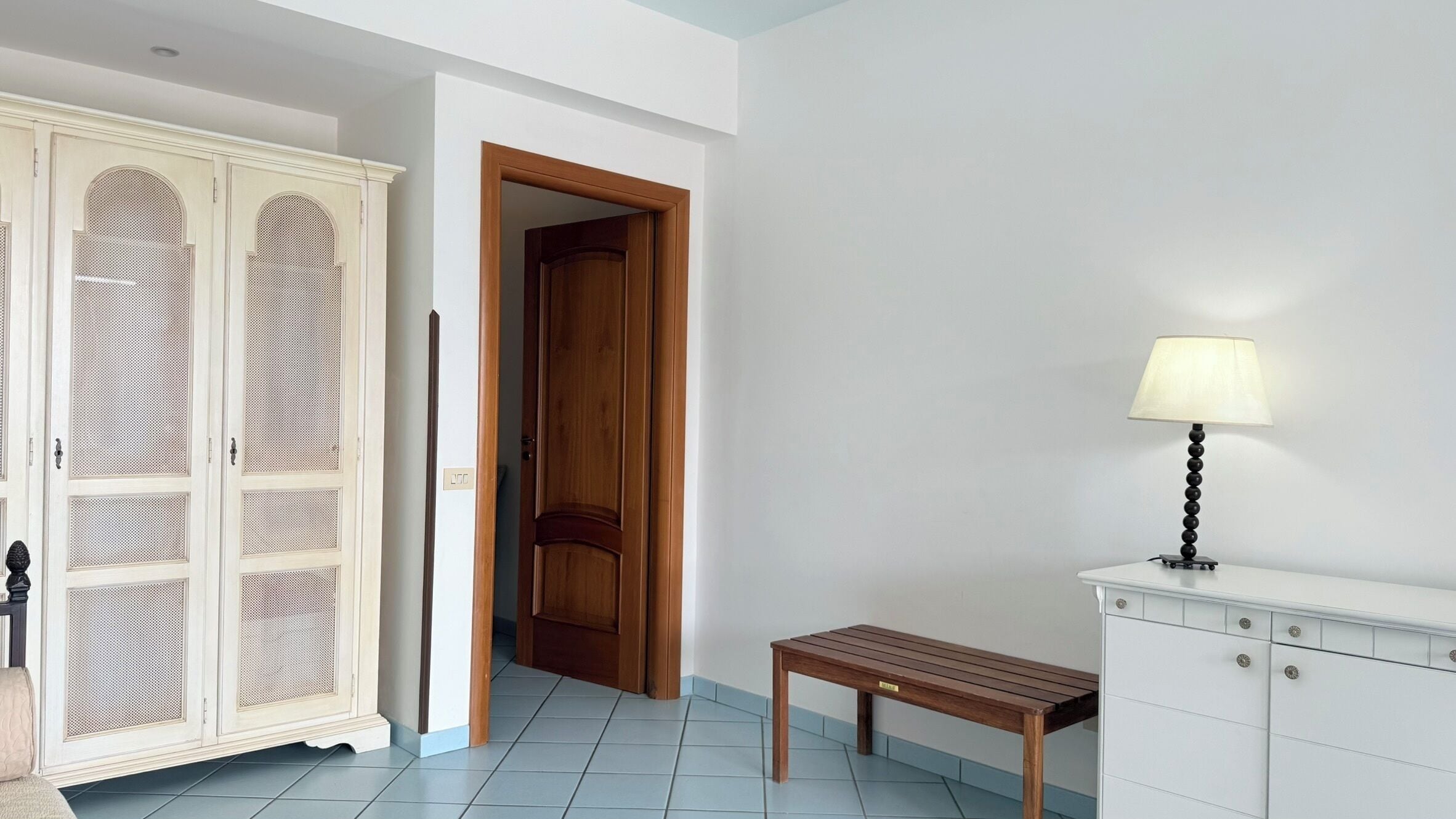 Un pasillo con una puerta de madera, un armario blanco con cajones, una pequeña mesa de madera y una lámpara de pie.