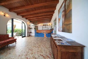 Una habitación amplia con techo de madera, piso de ladrillo, un sofá rojo, un escritorio de madera y un ventanal con vistas a zonas verdes.