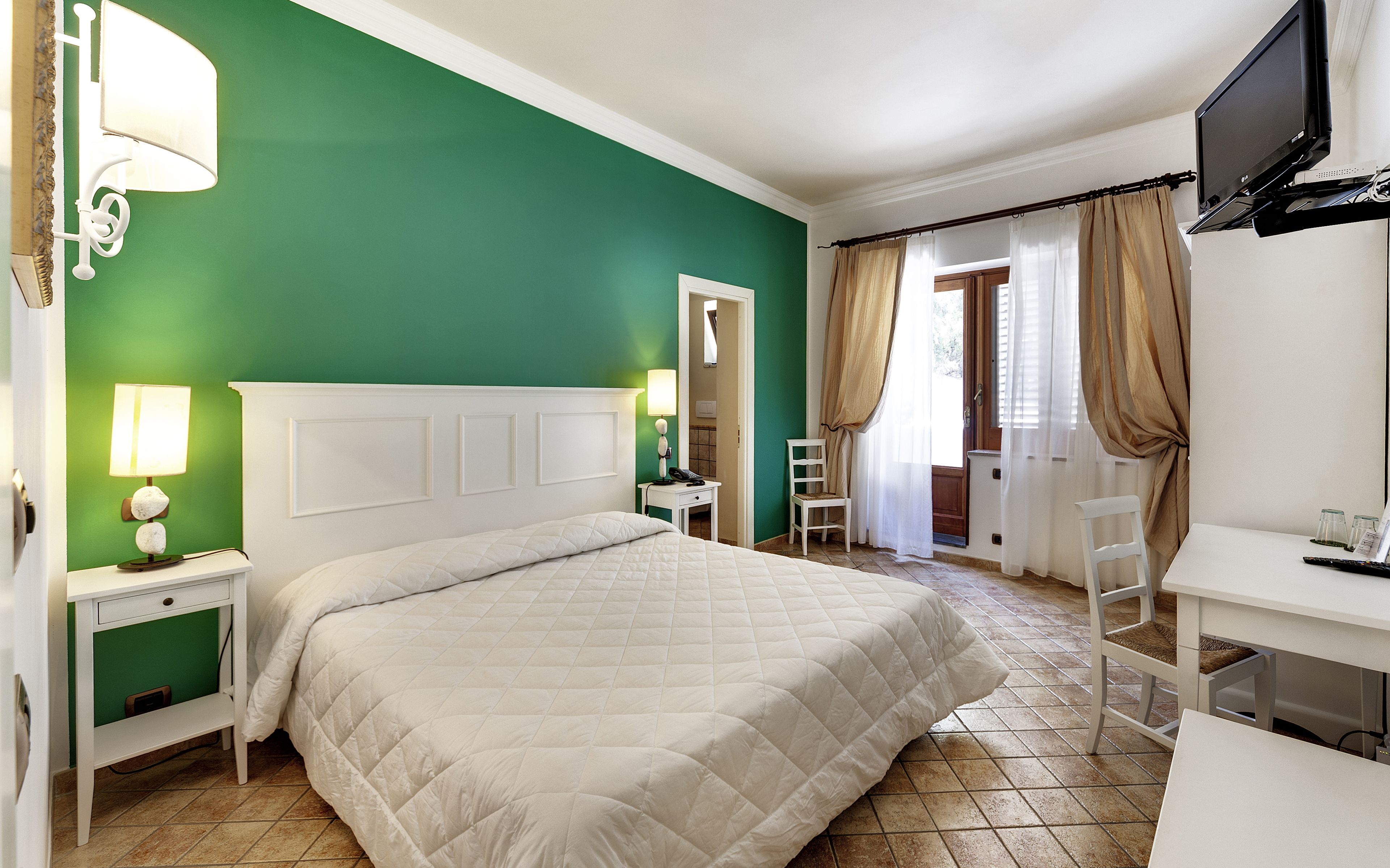 A hotel room with a large bed, a desk with a TV, a chair, and a lamp.