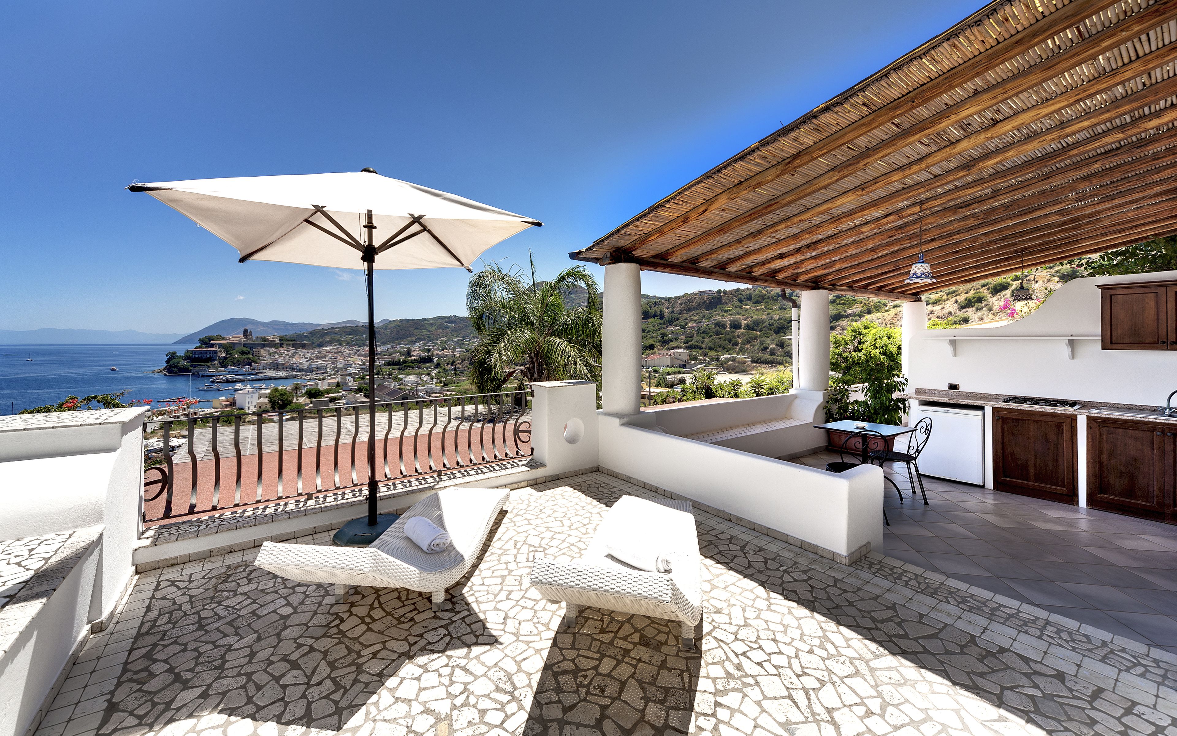 Eine Terrasse mit einem weißen Sonnenschirm, Korbsessel-Loungemöbeln, Steinplattenboden und Blick auf das Meer und die entfernten Hügel.