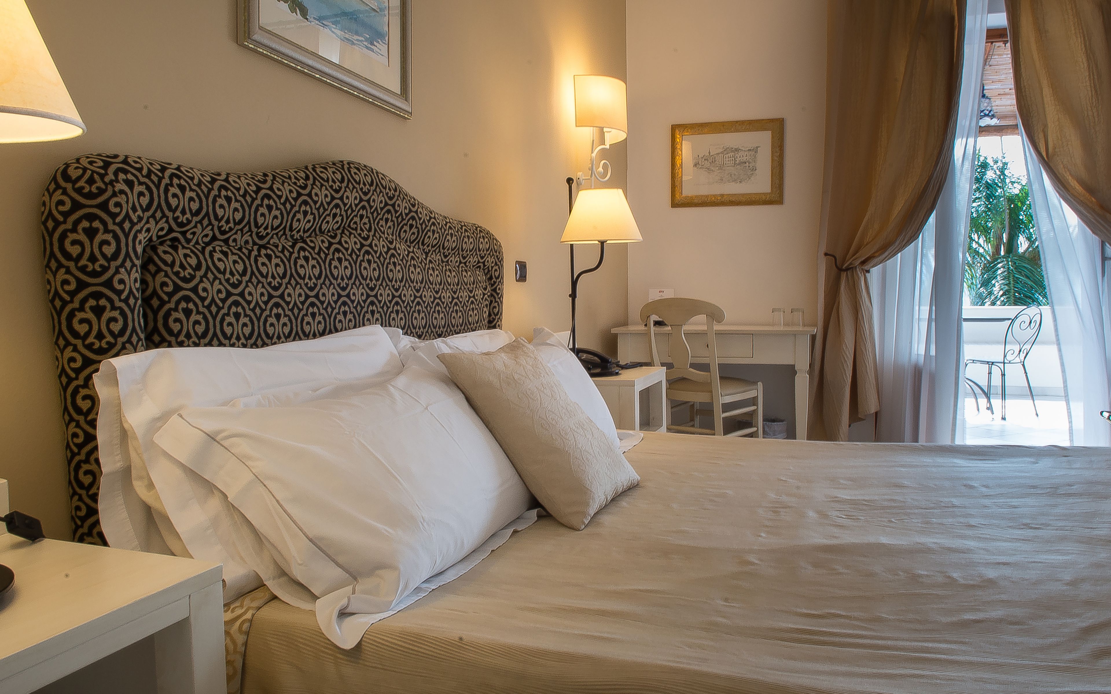 Une chambre d’hôtel avec une tête de lit à motifs, un bureau avec une chaise, une lampe et un lit recouvert de draps blancs.