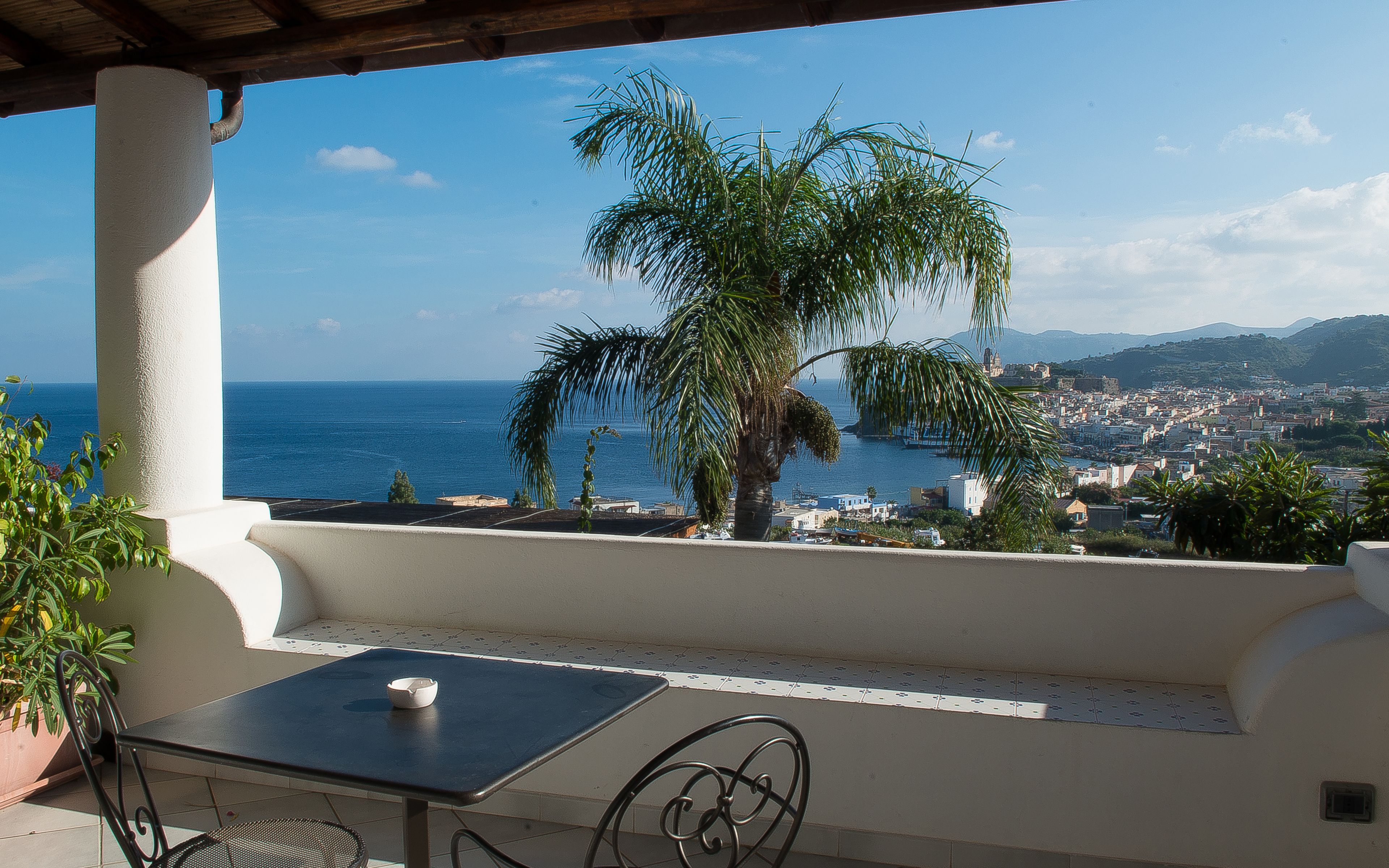 A balcony with a table and chairs, overlooking a coastal town and the sea.