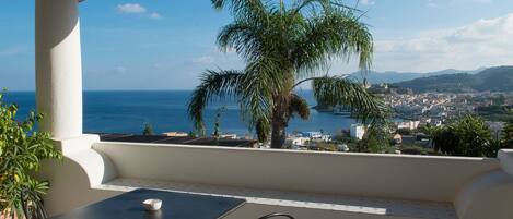 A balcony with a table and chairs, overlooking a coastal town and the sea.