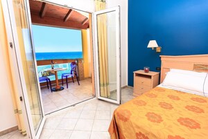 A hotel room with a bed, a nightstand, a table with chairs, and a view of the ocean through a sliding glass door.