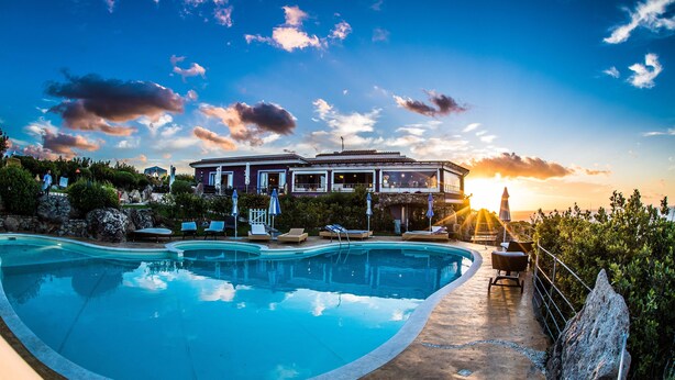 A swimming pool with lounge chairs and umbrellas, a house with a balcony overlooking the pool, and a sunset in the background.