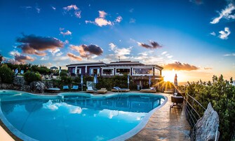 A swimming pool with lounge chairs and umbrellas, a house with a balcony overlooking the pool, and a sunset in the background.