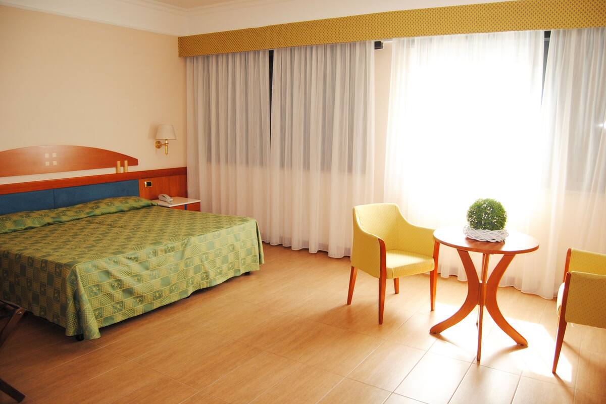 A hotel room with a bed, a desk, a chair, a small table with a plant, and a window with curtains.