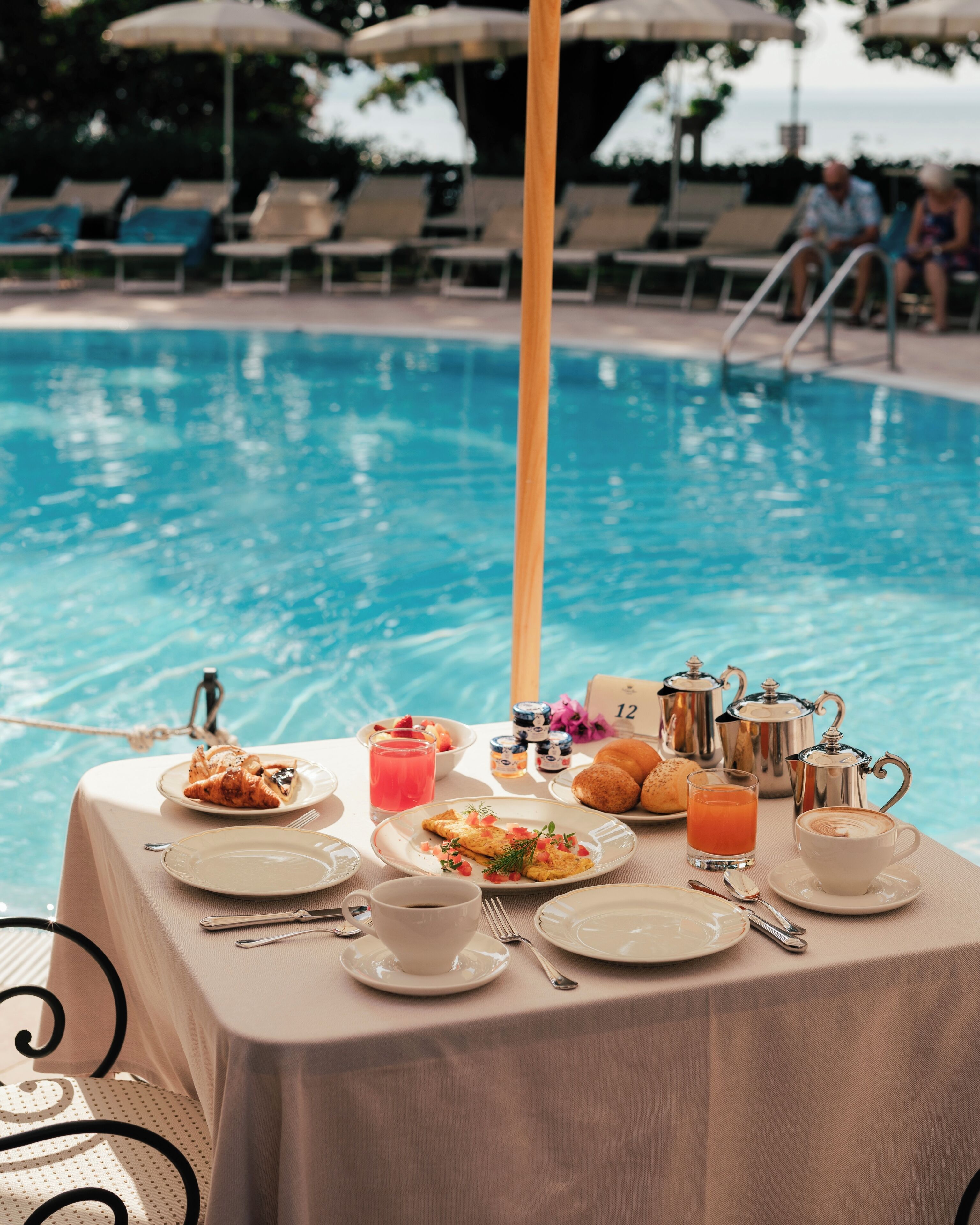 Ein Tisch, gedeckt für das Frühstück, neben einem Pool, mit Tellern, Tassen und einer Kaffeekanne.