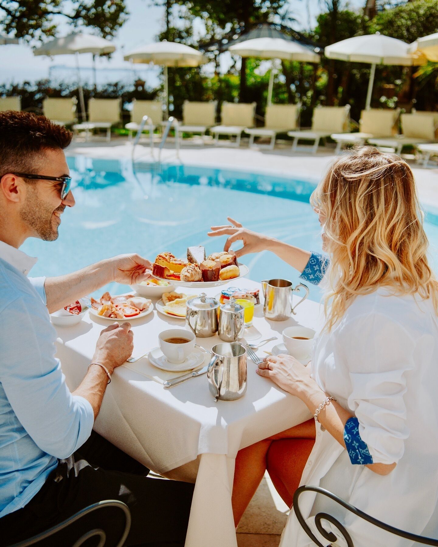 A couple dining by a poolside table with food, drinks, and coffee cups.