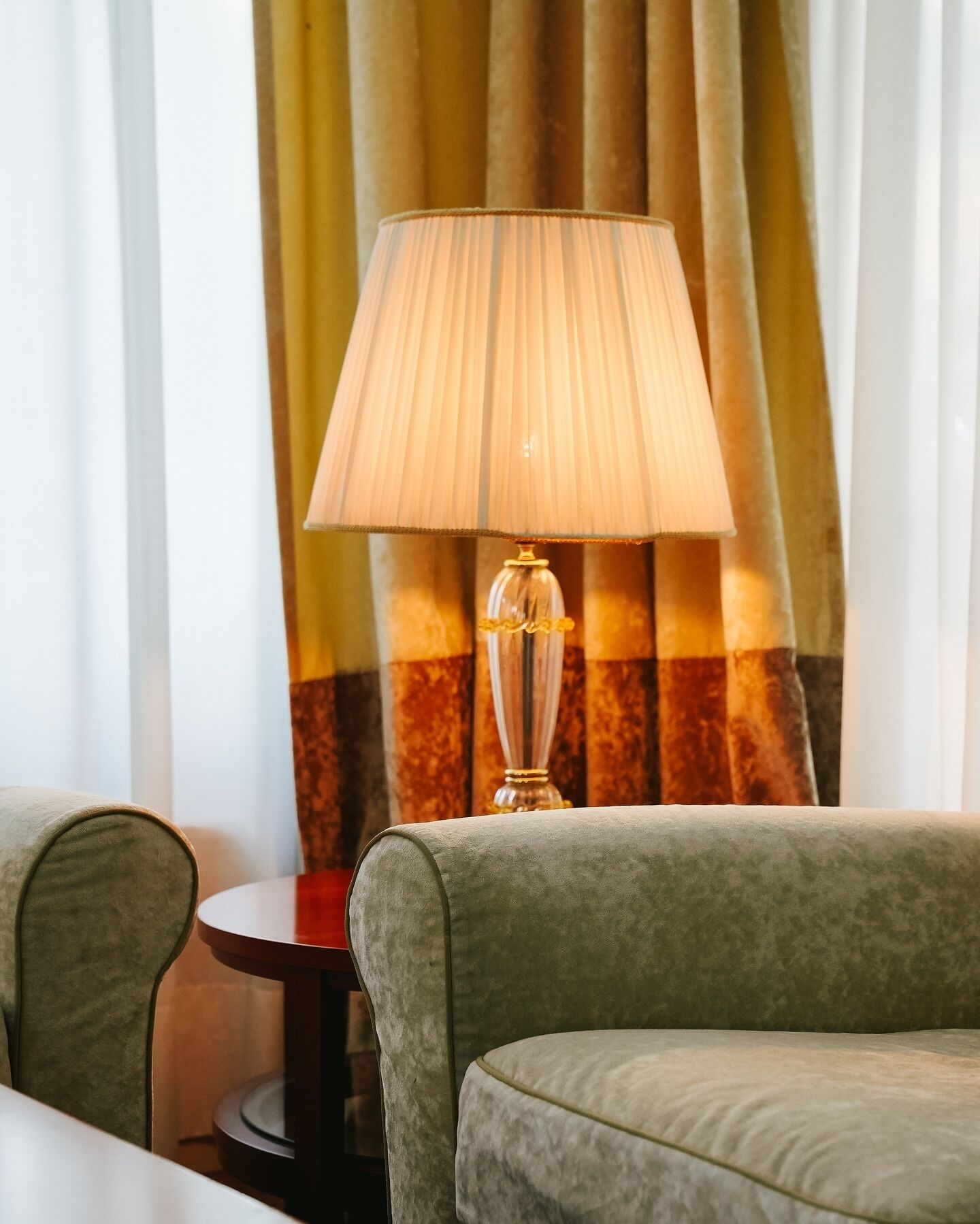 A hotel room with a green upholstered sofa, a small round table, and a lamp with a pleated lampshade.