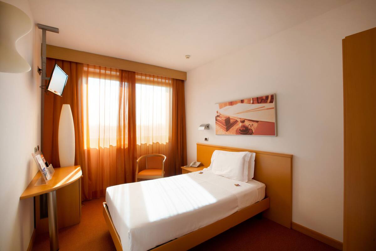 Una habitación de hotel con una cama, un escritorio con un monitor de computadora, una silla y un cuadro en la pared.