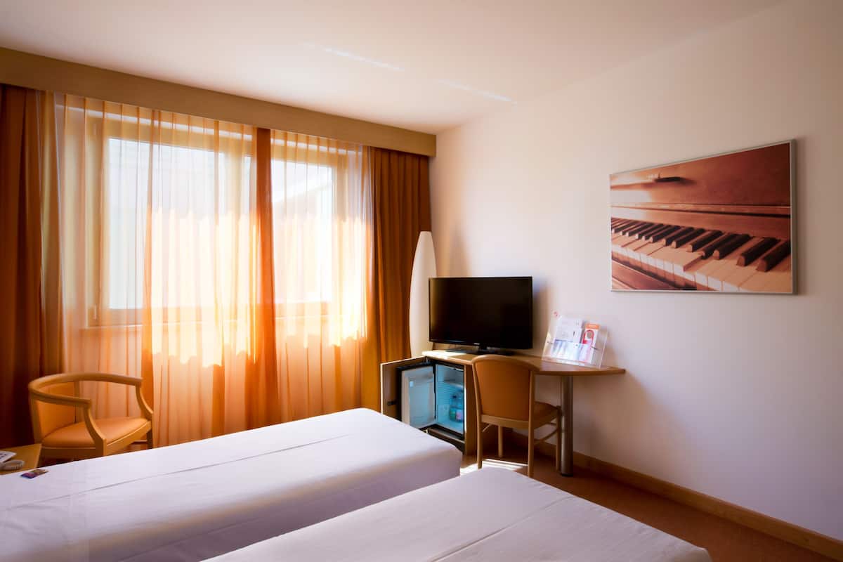 Habitación de hotel con dos camas, un escritorio con televisor, una silla y una obra de arte con temática de piano en la pared.