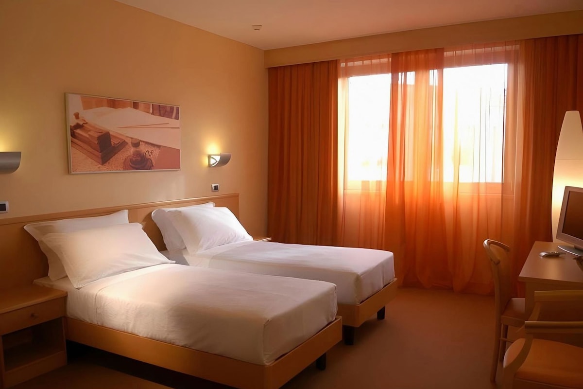 Habitación de hotel con dos camas, un escritorio con ordenador y un ventanal grande con cortinas.
