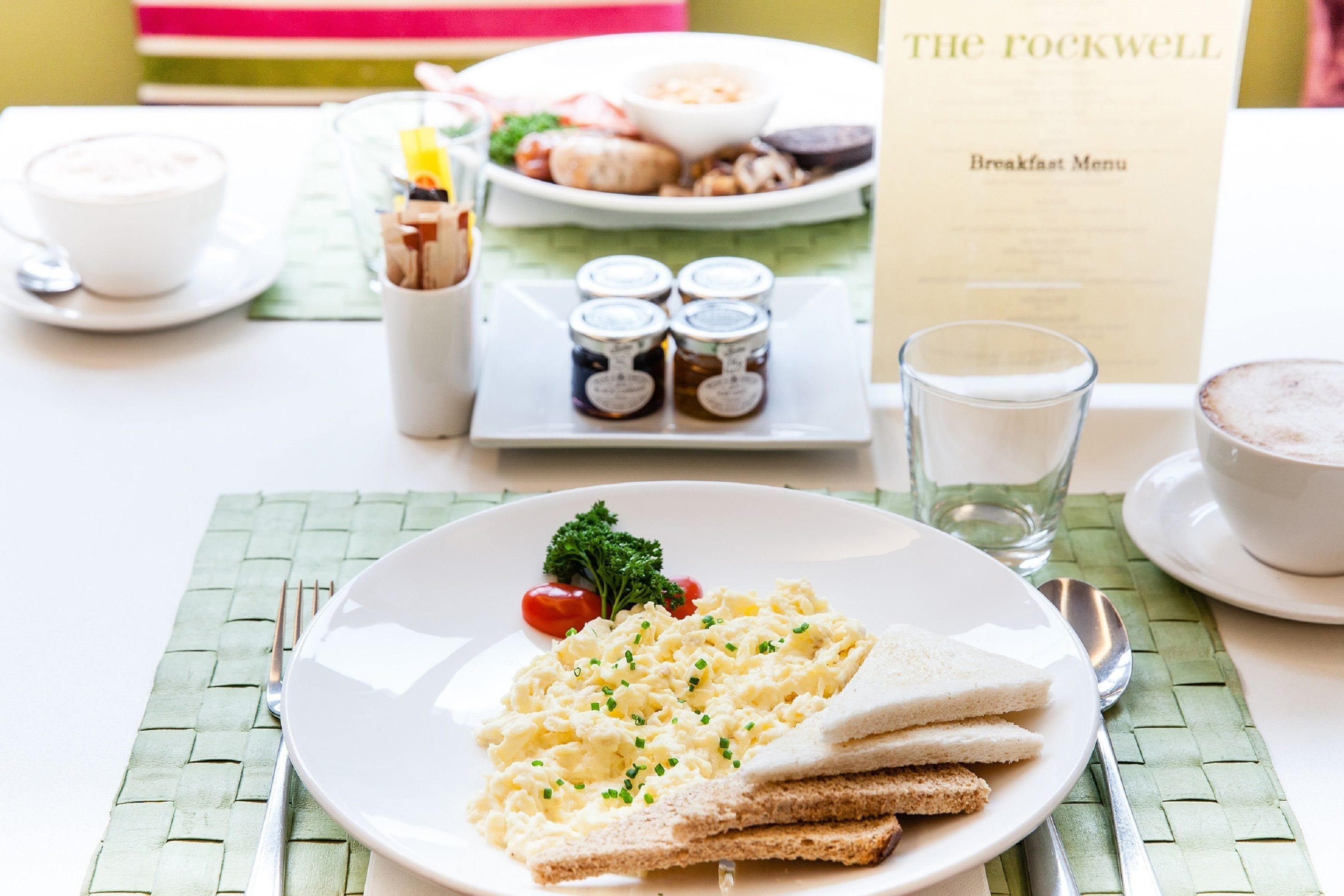 Un desayuno con huevos revueltos, tostadas y una guarnición de tomates en un plato blanco.
